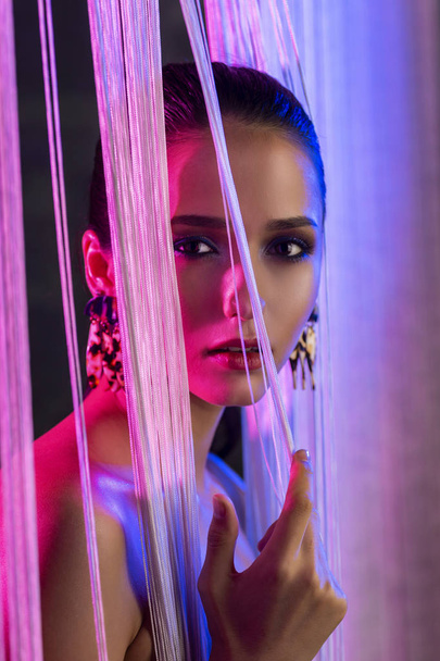 A beautiful glamorous brunette girl with naked shoulders wearing large elegant earrings peeps through a translucent curtain made of white threads. Multicolored artistic stage lighting. Copy space. - Foto, Bild