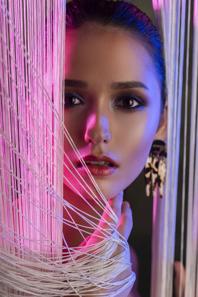 A beautiful glamorous brunette girl with naked shoulders wearing large elegant earrings peeps through a translucent curtain made of white threads. Multicolored artistic stage lighting. Copy space. - Foto, Imagen