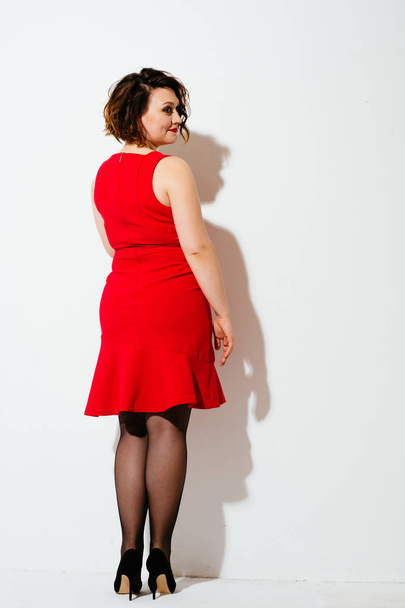 Visão traseira da linda senhora plus size em vestido vermelho e salto alto olhando sobre o ombro enquanto está em pé no fundo branco
 - Foto, Imagem