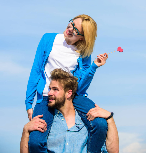 Romantic date concept. Man carries girlfriend on shoulders, sky background. Couple happy date having fun together. Woman holds heart on stick symbol of love. Couple in love walking outdoor sunny day - Photo, Image