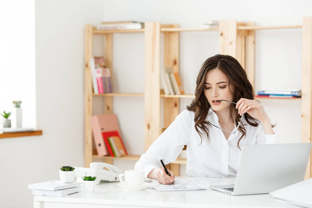 Молодая привлекательная женщина за современным офисным столом, работает с ноутбуком и думает о чем-то
. - Фото, изображение