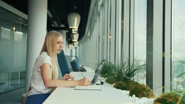 Businesswoman working with laptop in new office. Side view of woman sitting at table alongside window in modern office and using laptop in daylight. - Πλάνα, βίντεο