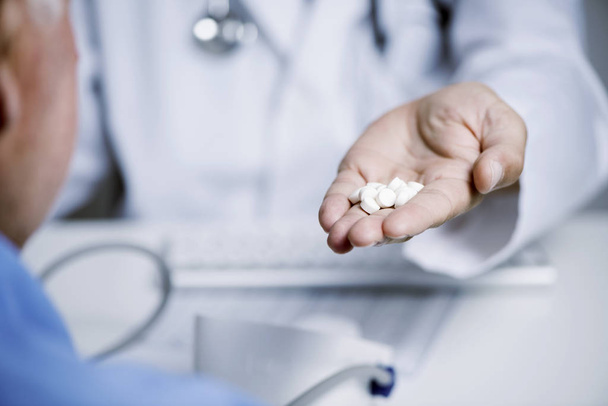 closeup ενός Καυκάσιος γιατρό άντρα, σε ένα λευκό παλτό, δίνοντας κάποια χάπια ένας ανώτερος άνθρωπος Καυκάσιων ασθενών, που κάθεται σε ένα γραφείο γιατροί - Φωτογραφία, εικόνα