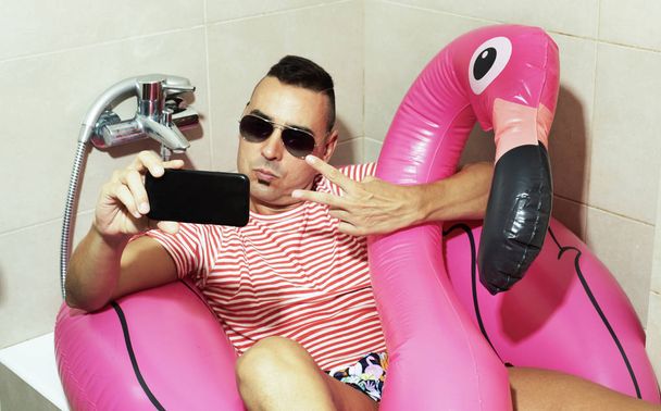 a young caucasian man wearing sunglasses, a swim suit and a swim ring in the shape of a pink flamingo in the bathtub of his bathroom, taking a self-portrait with his smartphone - Photo, Image
