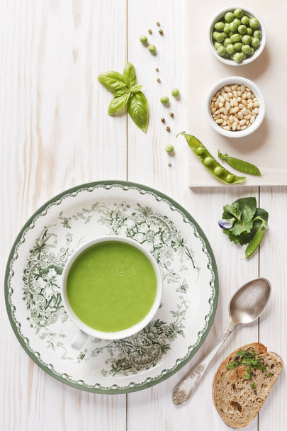Homemade fresh green pea cream soup with spices, pine nuts and toasted ciabatta bread. Close-up view from above on white wooden background - Photo, image