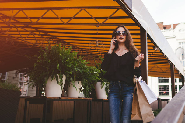 Kaunis tyylikäs nuori nainen ostoskasseineen kävelee ulos kahvilasta kaupungin kadulla ja puhuu puhelimessa. Muoti käsite
 - Valokuva, kuva