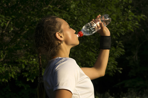 девушка после занятий спортом в парке утоляя жажду пьет воду из бутылки
 - Фото, изображение