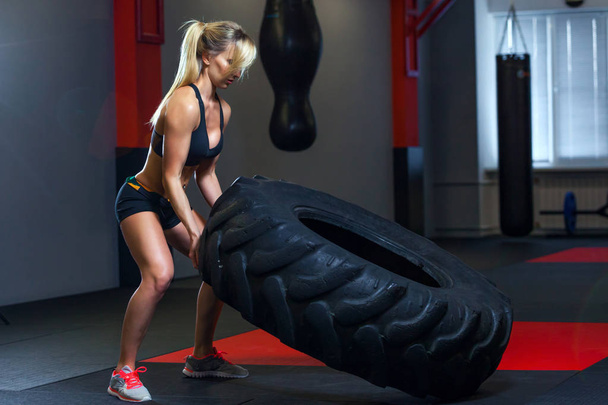 Fit female athlete working out with a huge tire, turning and carry in the gym. Crossfit woman exercising with big tire - Фото, изображение