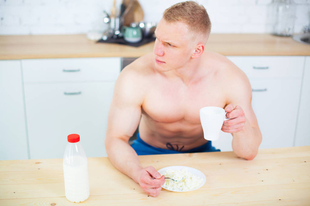 Muscular man with a naked torso in the kitchen with milk and cottage cheese, the concept of a healthy diet. Athletic way of life. - Photo, Image