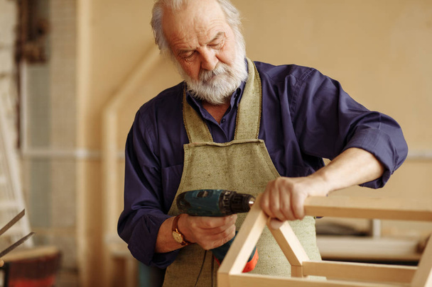 old grey headed man is working with drill in the home workshop - Foto, Bild