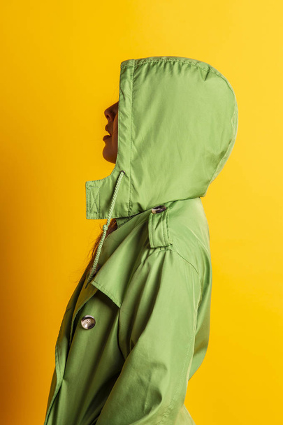 young pretty woman in green oilskin with hood posing against yellow background  - Φωτογραφία, εικόνα
