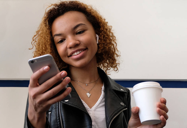 Egy gyönyörű fiatal modern fekete nő, a bőrkabát airpods a fülébe. A lány egy fehér üveg tart a kezében, a telefont, és mosolyog néz. - Fotó, kép