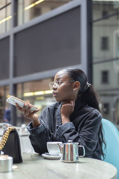atractiva mujer africana joven usando teléfono inteligente mientras toma una taza de té en la terraza
 - Foto, imagen