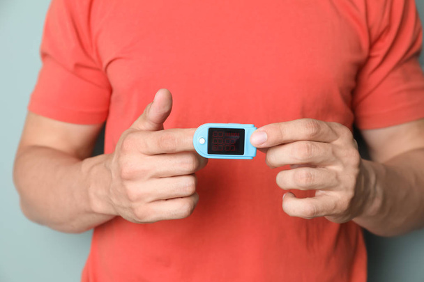 Young man checking pulse with blood pressure monitor on finger against color background, closeup - Fotó, kép
