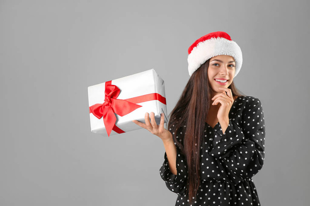Young beautiful woman in Santa hat with gift box on grey background. Christmas celebration - Фото, изображение