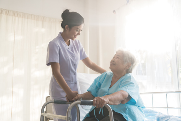 Ασιατικές νεαρά νοσοκόμα υποστηρίζοντας ηλικιωμένος ασθενής απενεργοποιημένη γυναίκα χρησιμοποιώντας walker στο νοσοκομείο. Έννοια της φροντίδας των ηλικιωμένων ασθενών - Φωτογραφία, εικόνα