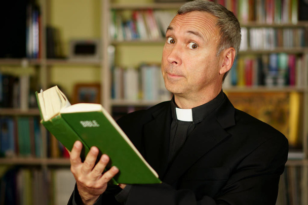 Um padre católico sério de boa aparência está estudando, lendo a Bíblia, em sua biblioteca. Ele olha para nós com desaprovação.
.                                - Foto, Imagem