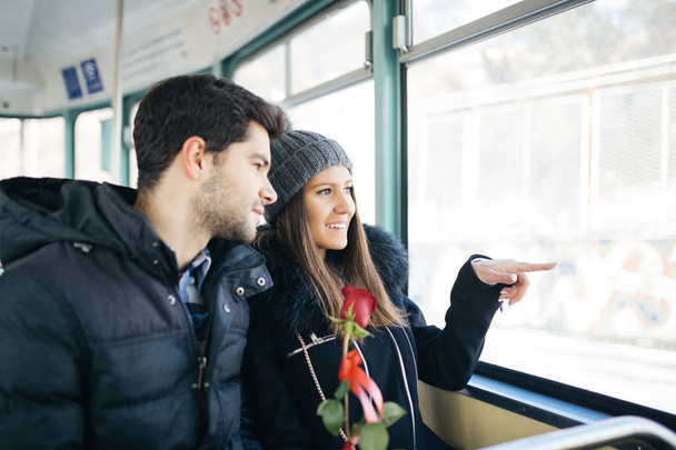 Красивая молодая пара влюблена, наслаждаясь вместе на трамвае, и мужчина дает красную косулю в качестве сюрприза для своей девушки
.  - Фото, изображение