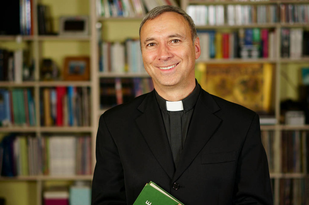 A good looking catholic priest is studying into his library. He looks at us with serenity and optimism.                                - Photo, Image