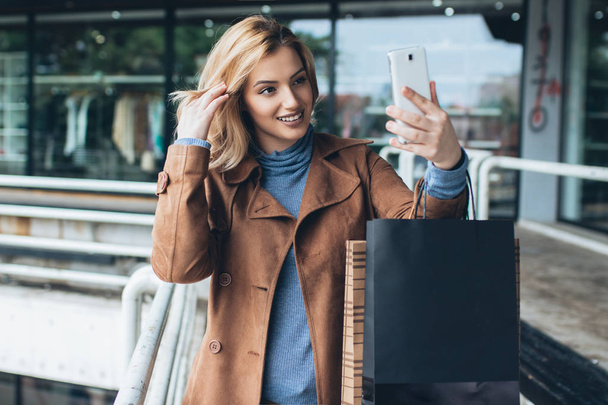 Bastante joven rubia tomando selfie mientras sostiene bolsas de compras
. - Foto, Imagen