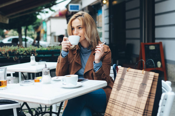 Mulher loira bonita sentada sozinha na cafetaria, fumando um cigarro e bebendo café
 - Foto, Imagem