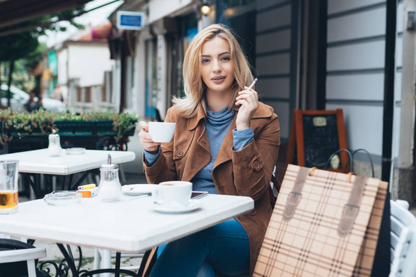 Mulher loira bonita sentada sozinha na cafetaria, fumando um cigarro e bebendo café
 - Foto, Imagem