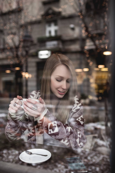 Красива червона голова молода жінка сидить в кафе ресторані, п'є капучино і дивиться на мобільний телефон
 - Фото, зображення
