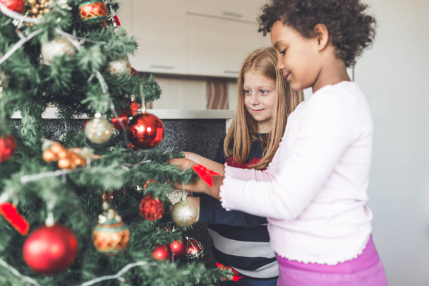 African and redhead little girls decorating the Christmas tree. Natural light.  - Foto, afbeelding