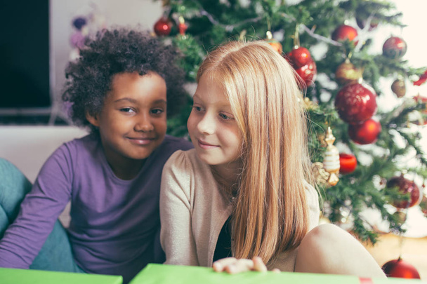 Αφρικής και κοκκινομάλλα κοριτσάκια με δώρα μαζί μπροστά από το χριστουγεννιάτικο δέντρο. Φυσικό φως.  - Φωτογραφία, εικόνα