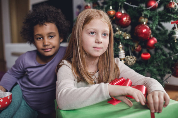 African and redhead little girls with gifts together in front of the Christmas tree. Natural light.  - 写真・画像
