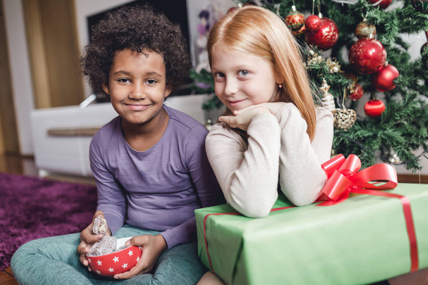 Niñas africanas y pelirrojas con regalos juntos frente al árbol de Navidad. Luz natural
.  - Foto, imagen