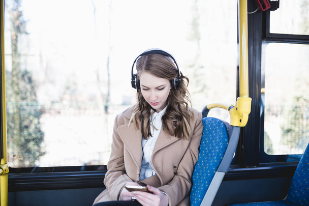 Όμορφη νεαρή ευτυχισμένη γυναίκα κάθεται σε αστικό λεωφορείο, βλέπουν το κινητό τηλέφωνο και να ακούτε μουσική.   - Φωτογραφία, εικόνα