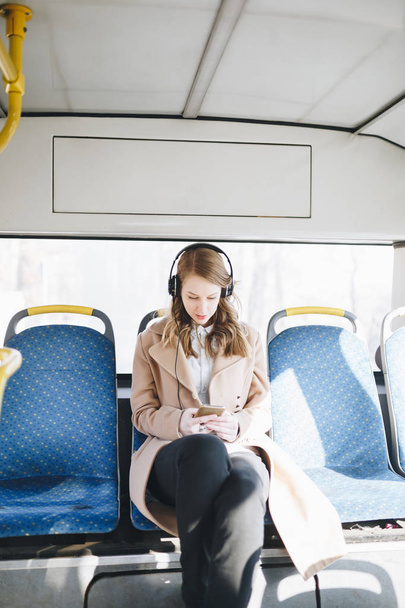 Beautiful happy young woman sitting in city bus, looking at mobile phone and listening to music.   - Foto, imagen