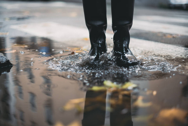 Woman in black rubber boots standing in a puddle with autumn leaves while it's raining - Photo, Image
