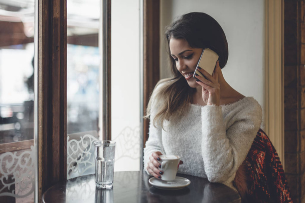 Hermosa mujer sentada sola en la cafetería, tomando café y hablando por teléfono celular
. - Foto, Imagen