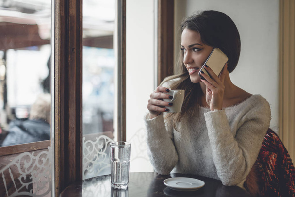 Beautiful woman sitting alone in cafeteria, drinking coffee and talking on cell phone. Selective focus on hand. - 写真・画像