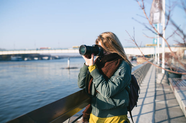 Jovem mulher em pé junto ao rio e tirando fotos com sua câmera. Belo dia ensolarado
. - Foto, Imagem