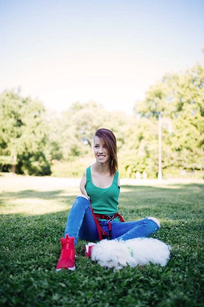 Retrato de una hermosa joven inválida sonriente nacida sin extremidades superiores disfrutando con su cachorrito blanco en el parque
.  - Foto, imagen
