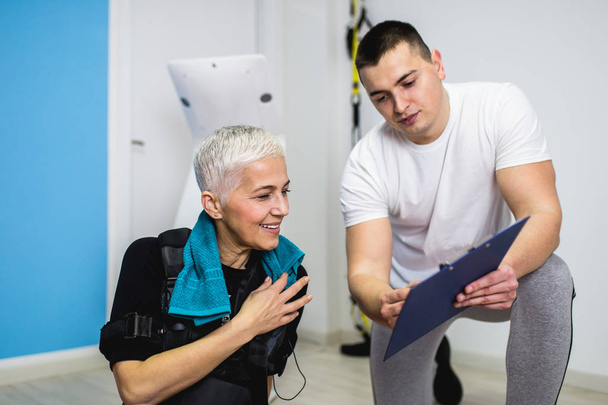 Belle femme âgée faisant des exercices en costume de stimulation musculaire électrique avec son entraîneur personnel au centre de réadaptation
 - Photo, image