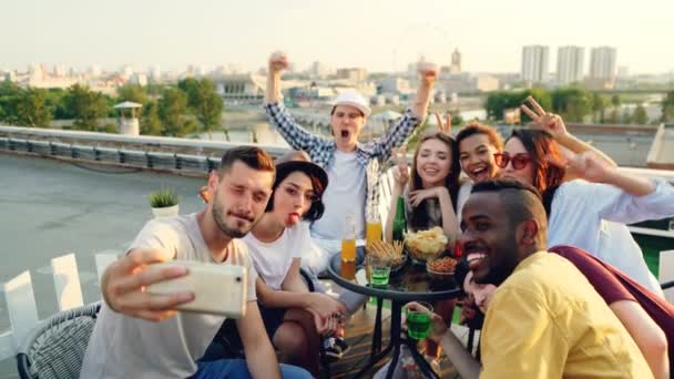 El tipo barbudo sostiene el teléfono inteligente y se toma selfies con amigos que posan con caras divertidas y gestos de manos sentados en la mesa en la azotea divirtiéndose.
. - Metraje, vídeo