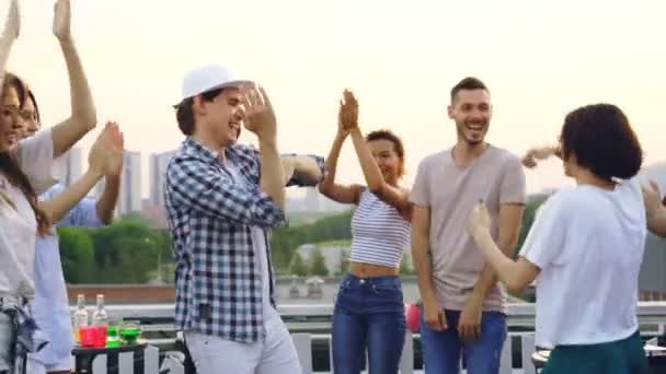 Bello giovane in berretto moderno sta ballando con gli amici alla festa sul tetto, uomini e donne si muovono e applaudono mani godendo l'estate e il tempo libero
. - Filmati, video