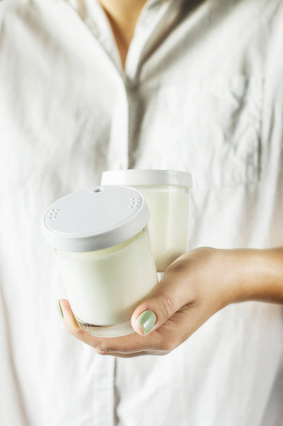Δύο γυάλινα βάζα με σπιτικό γιαούρτι στα χέρια της μια γυναίκα σε ένα λευκό πουκάμισο. Υγιεινή διατροφή πρωινό - Φωτογραφία, εικόνα