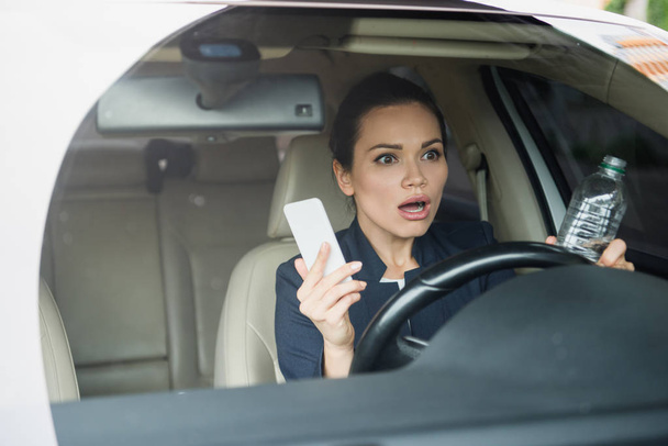shocked attractive woman holding smartphone and bottle of water while driving car - Foto, Imagen