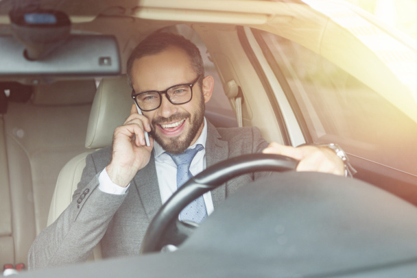 smiling handsome businessman driving car and talking by smartphone - 写真・画像