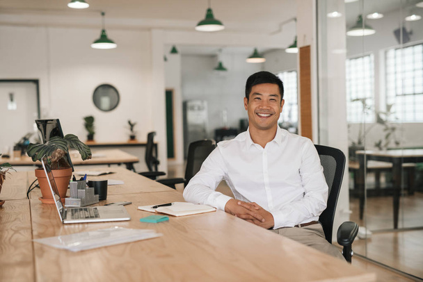 Porträt eines jungen asiatischen Geschäftsmannes, der mit einem Laptop lächelt und Notizen aufschreibt, während er an seinem Schreibtisch in einem modernen Büro sitzt - Foto, Bild