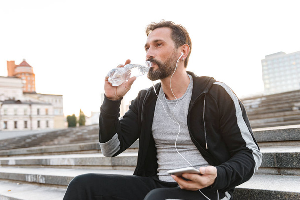 Attractive sportsman in earphones holding mobile phone while sitting on steps and drinking water from a bottle outdoors - Foto, Imagen