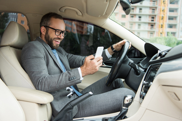 hombre sonriente guapo usando teléfono inteligente mientras conduce el coche
  - Foto, Imagen