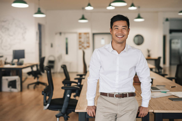 Smiling young Asian businessman leaning against a table while working alone in a large modern office - Valokuva, kuva