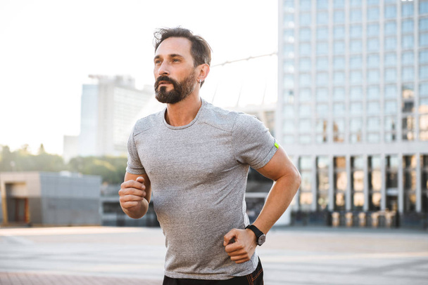 Image of handsome strong mature sportsman running at the street outdoors looking aside. - Photo, image