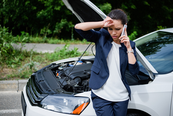 tired businesswoman talking by smartphone near broken car with open hood - Photo, Image